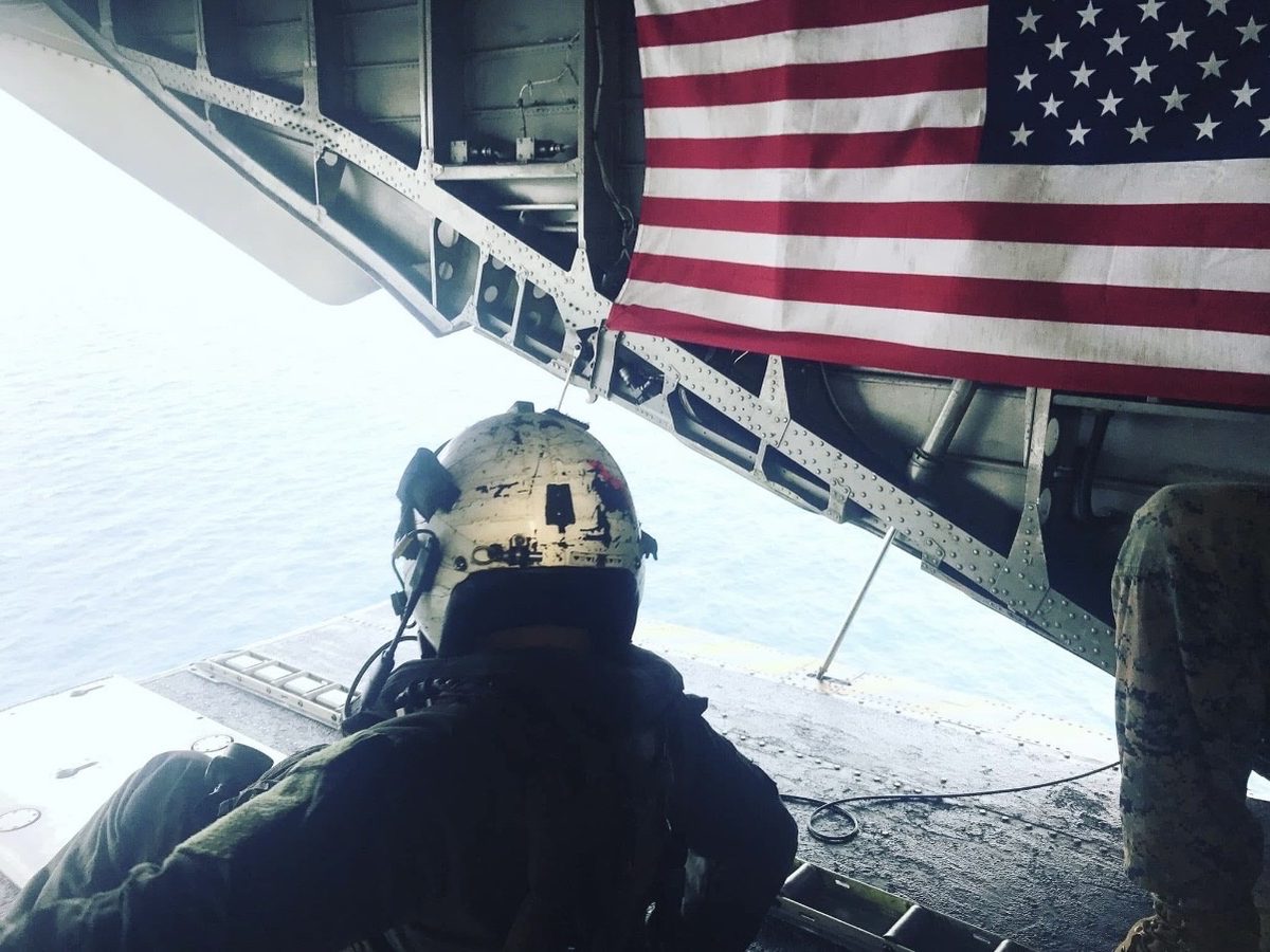 The view from an open aircraft ramp over bright open ocean, with an American flag hanging from the ramp overhead and a crew member seated in the doorway.