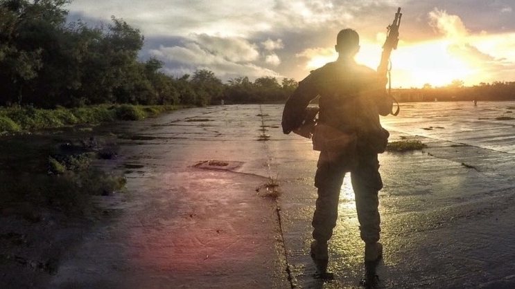 A Marine standing alone on a rain-soaked airstrip at sunset, rifle slung over one shoulder, the sky and standing water lit orange behind him.