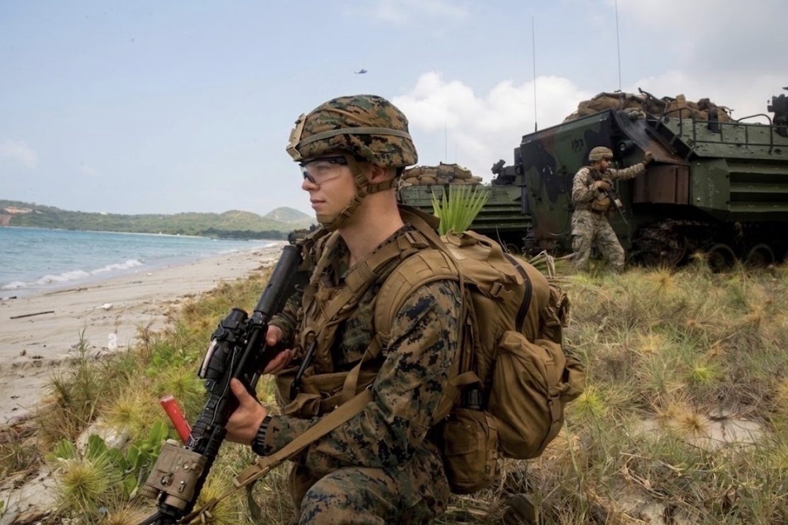 A Marine on a tropical beach with his rifle at the low ready, an amphibious assault vehicle and a second Marine in the dunes behind him.