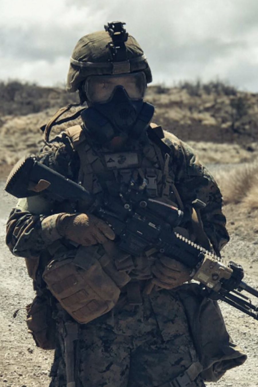 A Marine in full kit and a gas mask carrying a rifle along a desert road under a heavy overcast sky.