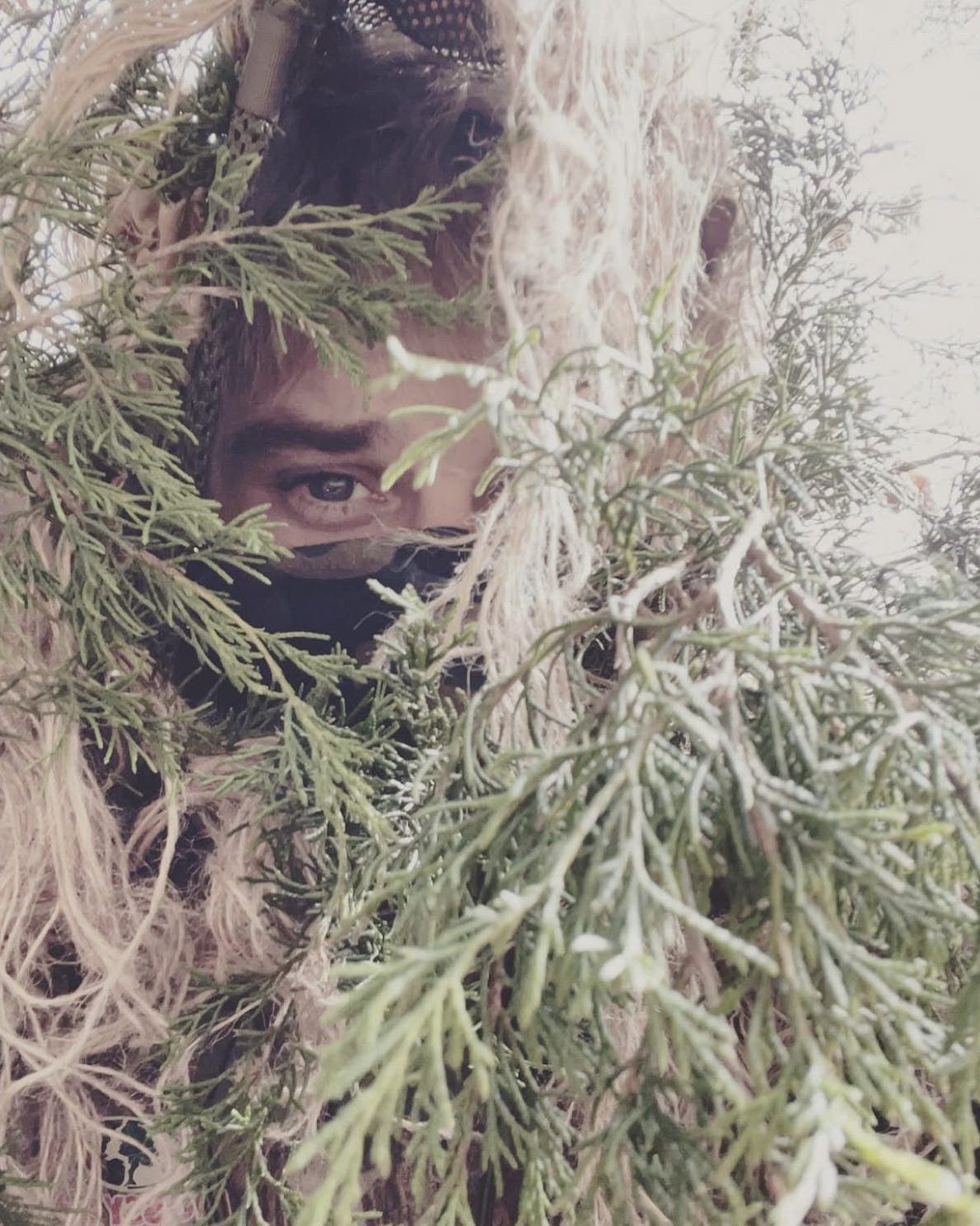 A close view of a Marine in a ghillie suit built from natural foliage, with only one eye visible through the netting.