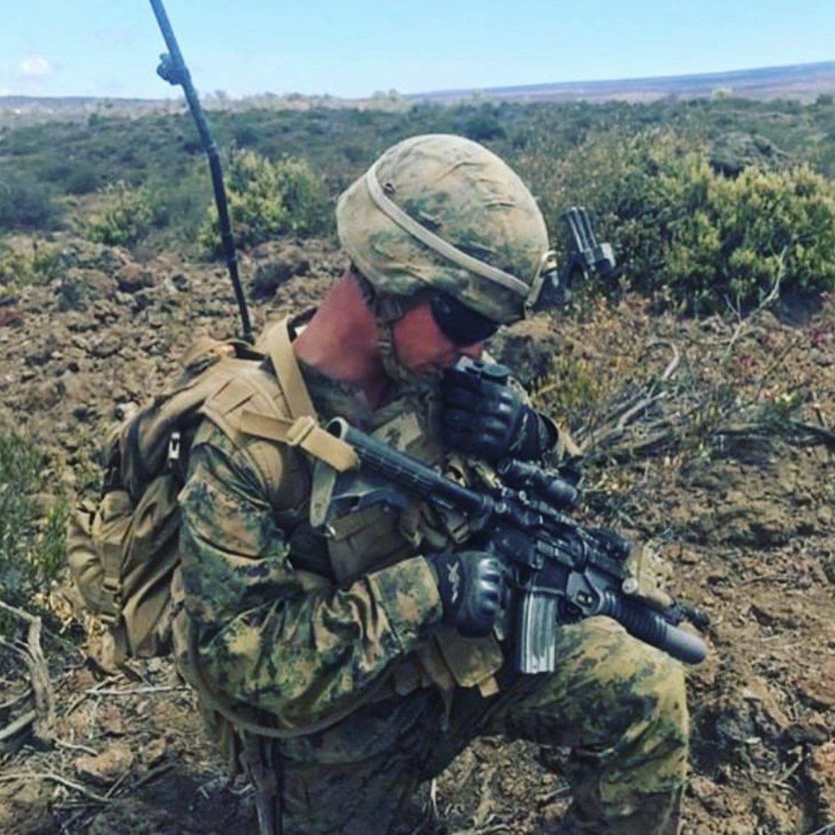 A Marine kneeling in low scrub with a radio antenna above him, speaking into a handset with his rifle braced across one knee.