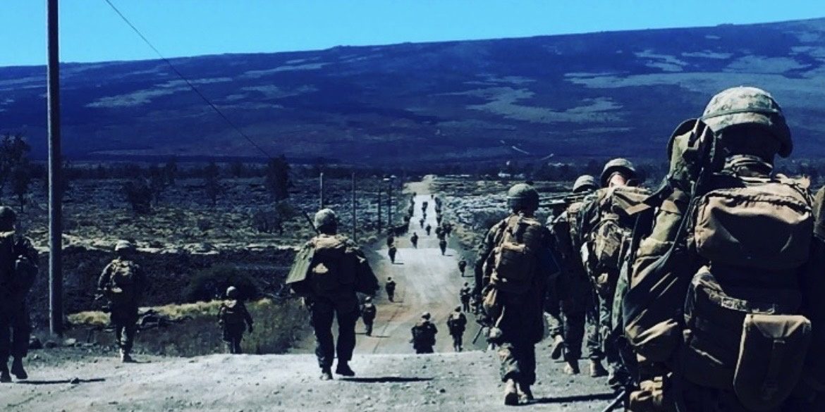 A patrol column of Marines walking a dirt road in single file, with a long dark mountain ridge in the distance.