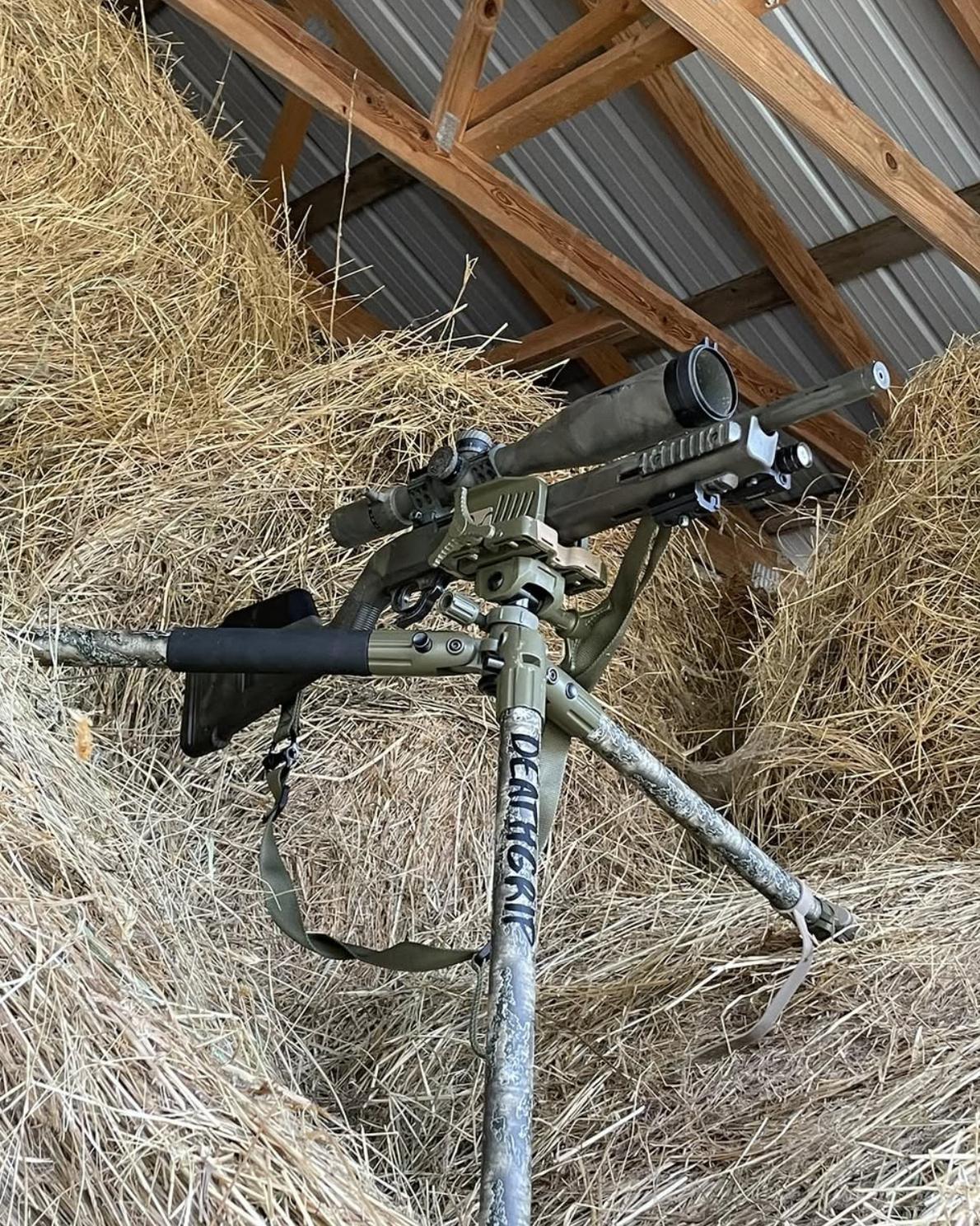 A precision rifle with a magnified optic mounted on a tripod, set up against stacked hay bales inside an open barn.