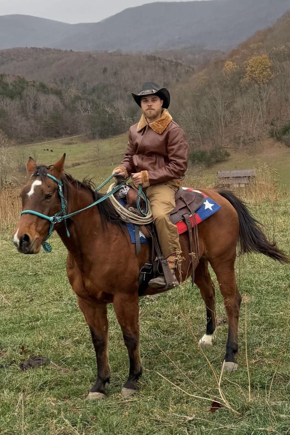 Ryder Bailey on horseback in a mountain pasture, wearing a cowboy hat and shearling-collared leather jacket, with Appalachian ridgelines and grazing cattle behind him.