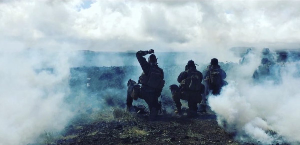 A team of Marines silhouetted in drifting white smoke on an open ridge, one signalling with a raised arm.