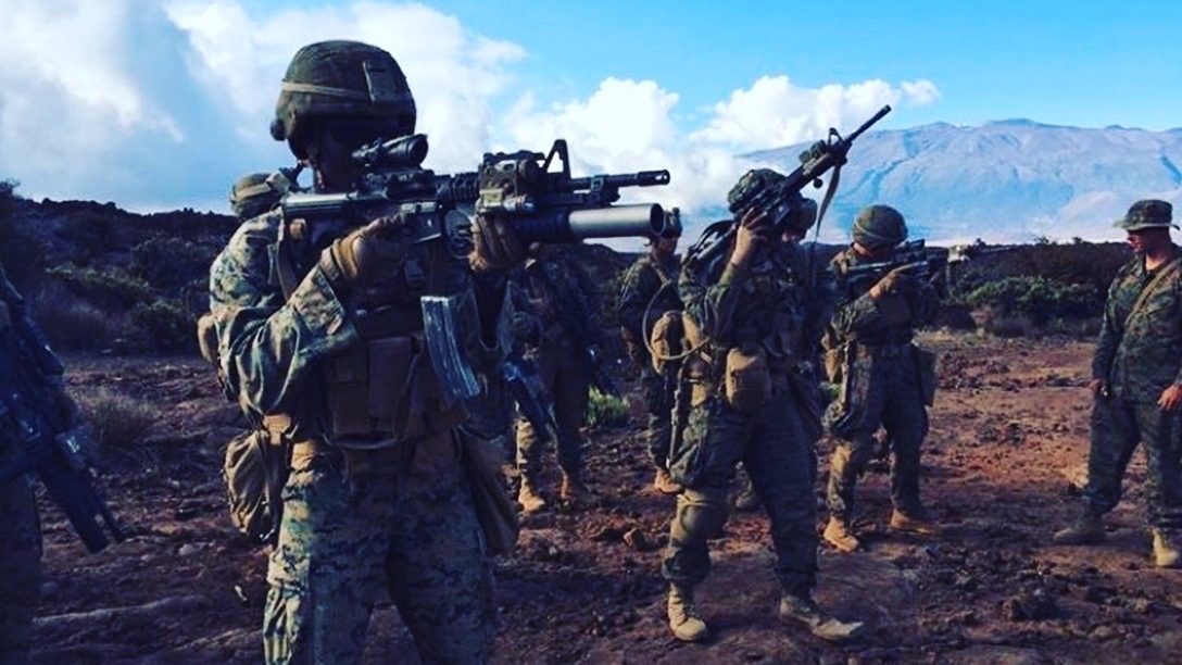 A squad of Marines spread across open volcanic ground with weapons shouldered, a broad mountain ridge and cloud bank behind them.