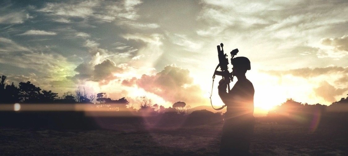 A Marine silhouetted against a wide sunset sky, rifle held upright, the sun flaring behind a distant treeline.